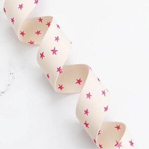 May include: Cream-coloured ribbon featuring a repeating pattern of bright pink stars. The ribbon is curled, and appears to be made of fabric. The background is white.