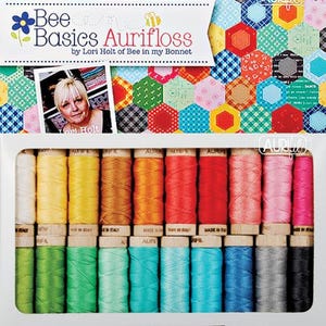 Könnte beinhalten: Ein Set mit 24 Spulen Aurifloss-Stickgarn in verschiedenen Farben, angeordnet in zwei Reihen in einer weißen Box. Die Box trägt ein Etikett mit der Aufschrift "Bee Basics Aurifloss by Lori Holt of Bee in my Bonnet".