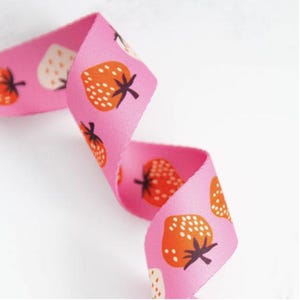 May include: A pink fabric ribbon featuring a repeating pattern of orange strawberries with white seeds and dark brown stems. The ribbon is curled into an S-shape, displaying the vibrant fruit print across its textured, woven surface.