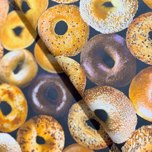 May include: A close-up of a fabric with a repeating pattern of various types of bagels. The bagels are in shades of brown, yellow, and white, and some have sesame seeds or poppy seeds on top.