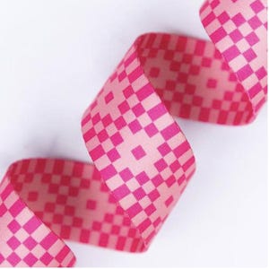 May include: A length of pink ribbon with a checkered pattern in a darker shade of pink. The ribbon is curled and appears to be made of fabric. The background is white.