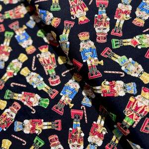 May include: Black fabric with a repeating pattern of colorful nutcracker figurines. The nutcrackers are in various poses and have gold accents.