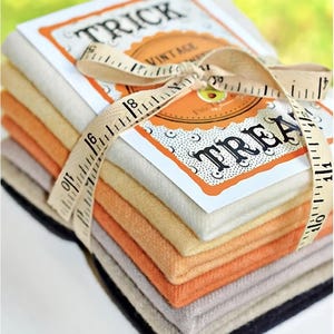 Op de afbeelding: Een stapel stofvierkanten in verschillende tinten oranje, beige en zwart, gebonden met een bruin lint en een meetlint. Het lint is gebonden in een strik en het meetlint is om de stapel gewikkeld. Een kaart met de woorden "Trick or Treat" ligt bovenop de stapel.