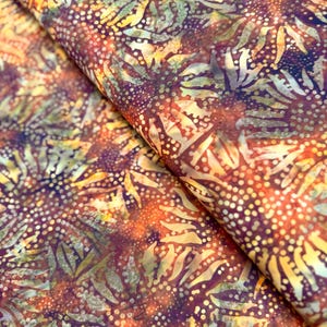 May include: Folded fabric with a batik pattern in shades of yellow, orange, and purple. The design includes a dotted pattern and leaf-like shapes. The fabric's folds reveal its texture and depth, creating a visually rich image.