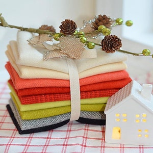 Op de afbeelding: Een stapel stofvierkanten in verschillende kleuren, waaronder wit, crème, oranje, geel, groen, grijs en zwart, gebonden met een wit lint. De stofvierkanten zijn in een stapel gerangschikt en liggen op een rood-wit geruit oppervlak. Een wit keramiek huis met een verlicht raam bevindt zich op de voorgrond.