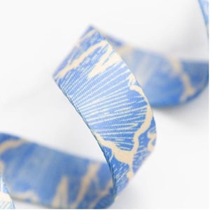 May include: Close-up of a blue ribbon with a cream-colored, abstract pattern. The ribbon is curled, showcasing the detailed design and texture. The ribbon's color is a light blue, with a cream-colored pattern.