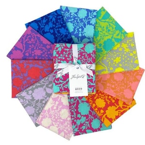 Pode incluir: Um conjunto de 10 quadrados de tecido com uma estampa floral em várias cores, incluindo rosa, azul, verde, amarelo e laranja. Os quadrados de tecido estão dispostos em círculo e amarrados com uma fita branca. O rótulo na fita diz "Free Spirit".