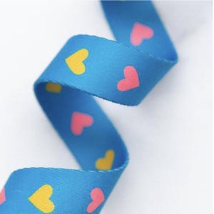 May include: A blue ribbon featuring a repeating pattern of yellow and pink hearts. The ribbon is curled, displaying the pattern on both sides. The hearts are evenly spaced across the ribbon's surface.