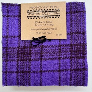 Op de afbeelding: Een vierkant stuk handgeverfde wollen stof in een paars en donkerpaars ruitpatroon. Een bruin label is bevestigd met zwart touw, met de tekst "HAND DYED WOOL FROM: Primitive Gatherings". Het label bevat ook contactgegevens.