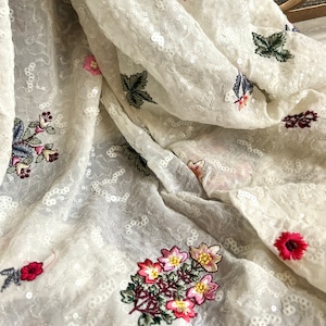 May include: White sheer fabric with embroidered floral designs in shades of pink, red, green, and yellow. The fabric is covered in small white sequins.