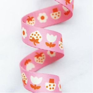 May include: A pink fabric ribbon featuring a repeating pattern of white and orange strawberries, flowers, and mushrooms. The ribbon is coiled in a spiral shape, displaying the printed design clearly across the smooth, woven material surface.