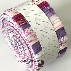 May include: A roll of fabric featuring a variety of floral prints in shades of pink, purple, and white. The fabric is wrapped around a white label with the text "CAMELOT FABRICS FRESHLY MADE" repeated multiple times.