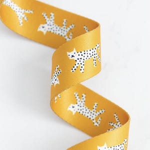 May include: Mustard yellow ribbon featuring a repeating pattern of white spotted cat illustrations. The ribbon is curled, displaying the design from various angles. Suitable for crafting or gift wrapping.