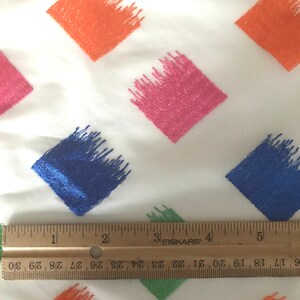 May include: A white fabric with a repeating pattern of colourful diamond shapes. The diamonds are filled with a brushstroke design in shades of orange, pink, blue, and green. A ruler is placed at the bottom of the image to show the scale.