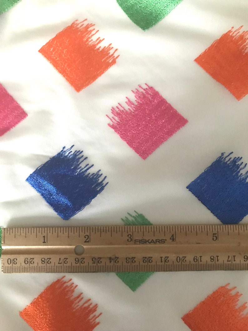 May include: A white fabric with a repeating pattern of colourful diamond shapes. The diamonds are filled with a brushstroke design in shades of orange, pink, blue, and green. A ruler is placed at the bottom of the image to show the scale.