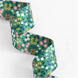 May include: A green ribbon with a floral design. The ribbon displays small flowers in pink, yellow, and white, accompanied by green leaves and stems. The ribbon is curled, forming a decorative shape. The background is white.