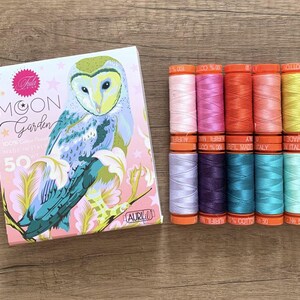 Op de afbeelding: Een doos met 50 spoelen 100% katoenen borduurgaren in verschillende kleuren, waaronder roze, paars, turquoise en geel. De doos heeft een fantasievolle illustratie van een uil met een maan en sterren. De tekst "Moon Garden" staat op de doos gedrukt.