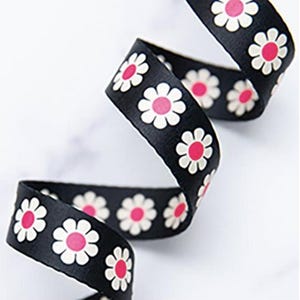 May include: Black ribbon featuring a repeating pattern of white daisies with pink centres. The ribbon is curled, suggesting a smooth, woven texture. The daisies are evenly spaced along the ribbon.