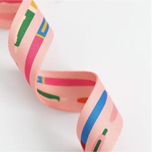 May include: A pink ribbon featuring a repeating pattern of colourful pencils. The pencils are in shades of green, blue, pink, and yellow. The ribbon is curled and appears to be made of fabric.