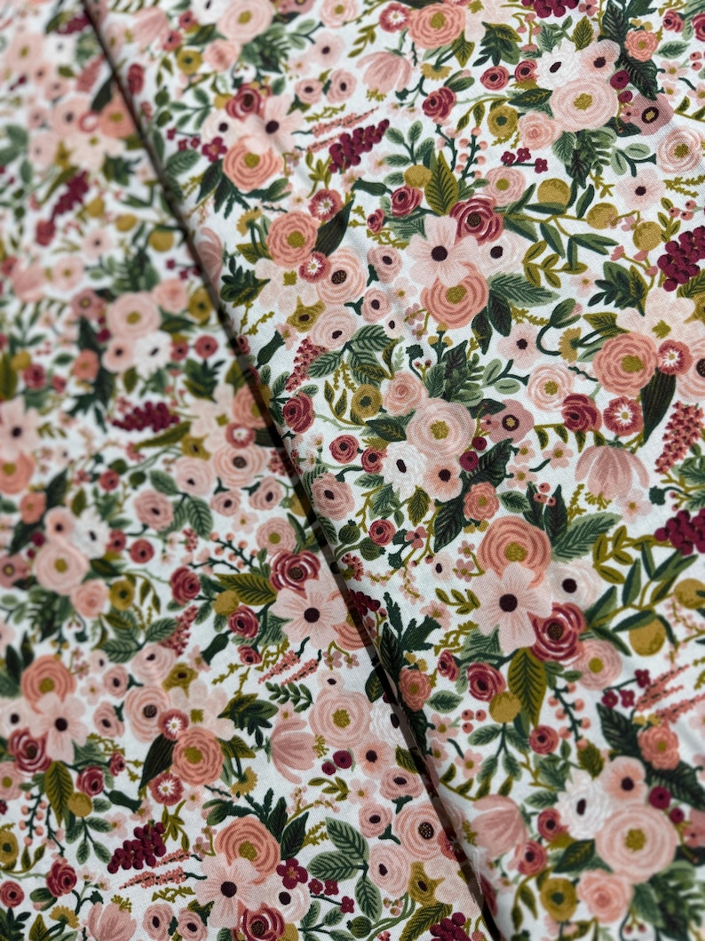 May include: A white fabric with a pink and green floral pattern. The fabric features a variety of flowers, including roses, daisies, and lilies. The flowers are arranged in a random pattern and are surrounded by green leaves.