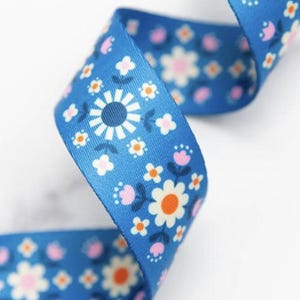 May include: Close-up of a blue ribbon with a floral design. The ribbon has white, orange, and pink flowers. The ribbon is curled and appears to be made of fabric. The background is white.