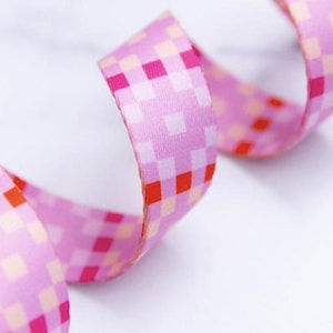 May include: A length of fabric ribbon featuring a repeating pixelated pattern in shades of light pink, magenta, and bright red. The ribbon is curled into a spiral shape, displaying the smooth texture and the consistent grid-like design.