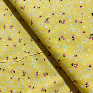 May include: Yellow fabric with a repeating pattern of small pink cherries, white flowers, and tiny green and white dots.