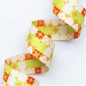 May include: A patterned ribbon in orange, green, and white, featuring geometric shapes like squares, circles, and flowers. The ribbon is curled, suggesting it's made of fabric. The design is repeated along the length.