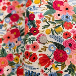 May include: A white fabric with a floral pattern featuring red, pink, blue, and yellow flowers. The fabric is covered in a variety of flowers and leaves.