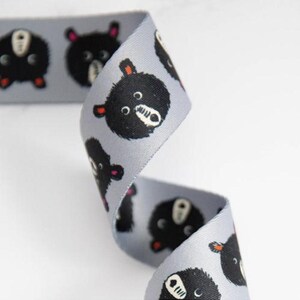 May include: A grey ribbon featuring a repeating pattern of black bear faces. The bears have white muzzles, small red ears, and black eyes. The ribbon is curled and appears to be made of a smooth, satin-like material.