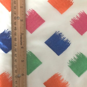 May include: A white fabric with a repeating pattern of colourful diamond shapes. The diamonds are outlined in white and filled with shades of green, blue, pink, and orange. A ruler is placed on the fabric to show the scale.