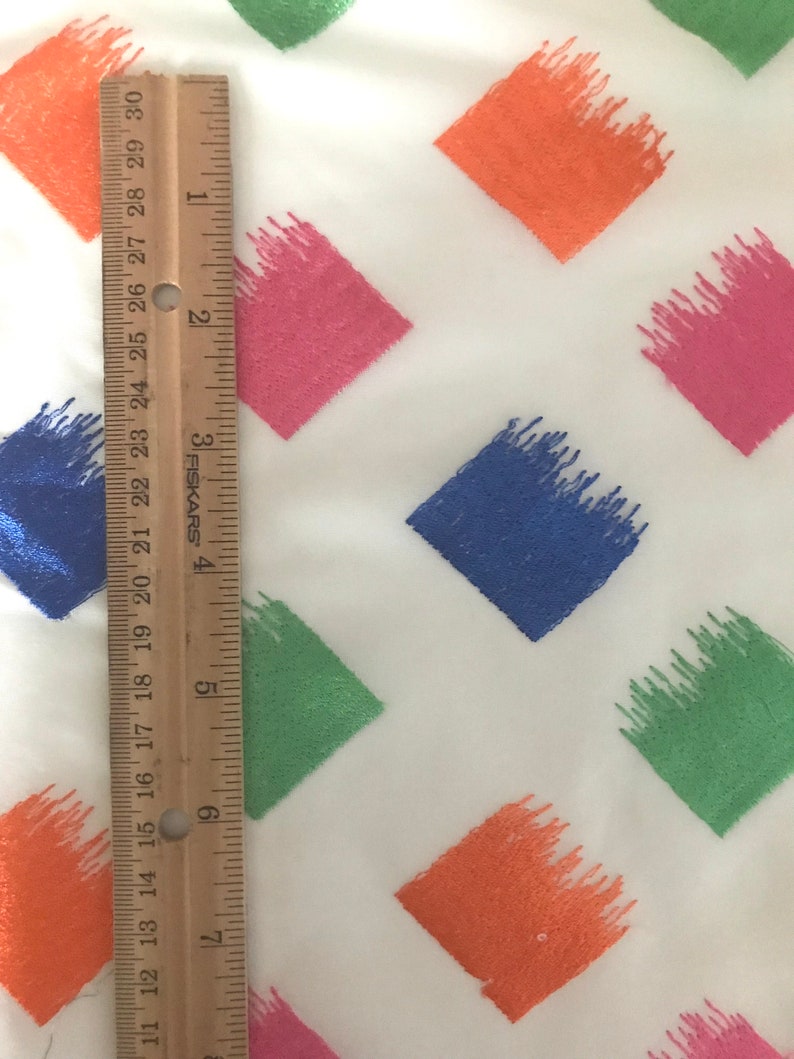 May include: A white fabric with a repeating pattern of colourful diamond shapes. The diamonds are outlined in white and filled with shades of green, blue, pink, and orange. A ruler is placed on the fabric to show the scale.