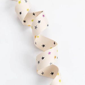 May include: Cream-coloured ribbon featuring a repeating pattern of small stars in yellow, black, grey, and pink. The ribbon is curled in a spiral shape. Plain white background.