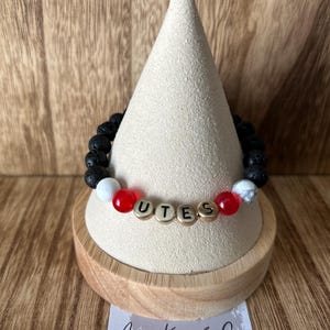 May include: A black lava rock bracelet with red and white beads and gold letter beads spelling "UTES". The bracelet is displayed on a wooden stand with a business card that reads "Asher Krew & Co."