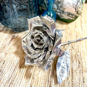 May include: A handcrafted silver metal rose with detailed petals and a stem. The rose has a leaf with the text "2011-2021" engraved on it. The rose is displayed on a textured surface.