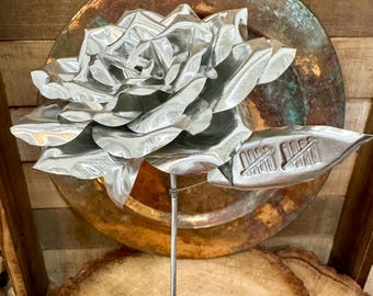 May include: A silver-colored metal rose sculpture with layered, folded petals and a single leaf attached to a thin wire stem. The leaf features a stamped tally mark design. The flower is crafted from reflective, smooth sheet metal material.