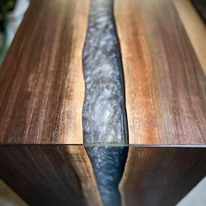 May include: A wooden table top with a natural wood grain finish. A thick, clear epoxy resin river runs down the center of the table, creating a unique and modern look.