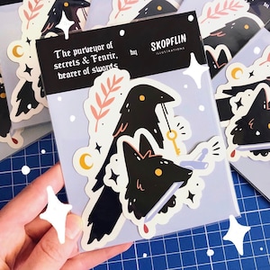 May include: A sticker featuring a black raven with a key and a wolf's head with a knife through it. The sticker has a light blue background with white stars and the text "The purveyor of secrets & Fenrir, bearer of swords" and "SKOPFLIN ILLUSTRATIONS".