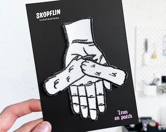 Op de afbeelding: Witte strijk-op patch met een ontwerp van drie handen die elkaar vasthouden. De patch is op een zwarte achtergrond met de tekst "SKOPFLIN ILLUSTRATIONS" en "Iron on patch".