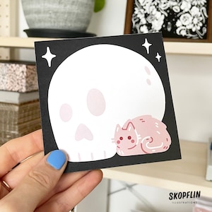 May include: A black and white sticker with a pink cat and a white skull with pink details. The sticker has a black background and white stars. The text "SKOPFFLIN ILLUSTRATIONS" is at the bottom of the sticker.