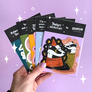 May include: A set of six sticker sheets featuring illustrations of badgers, mushrooms, and other woodland creatures. The sticker sheets are black with white text and are packaged in a clear plastic sleeve. The text on the sticker sheets reads "Badger stickerset by Skoplin Illustrations".
