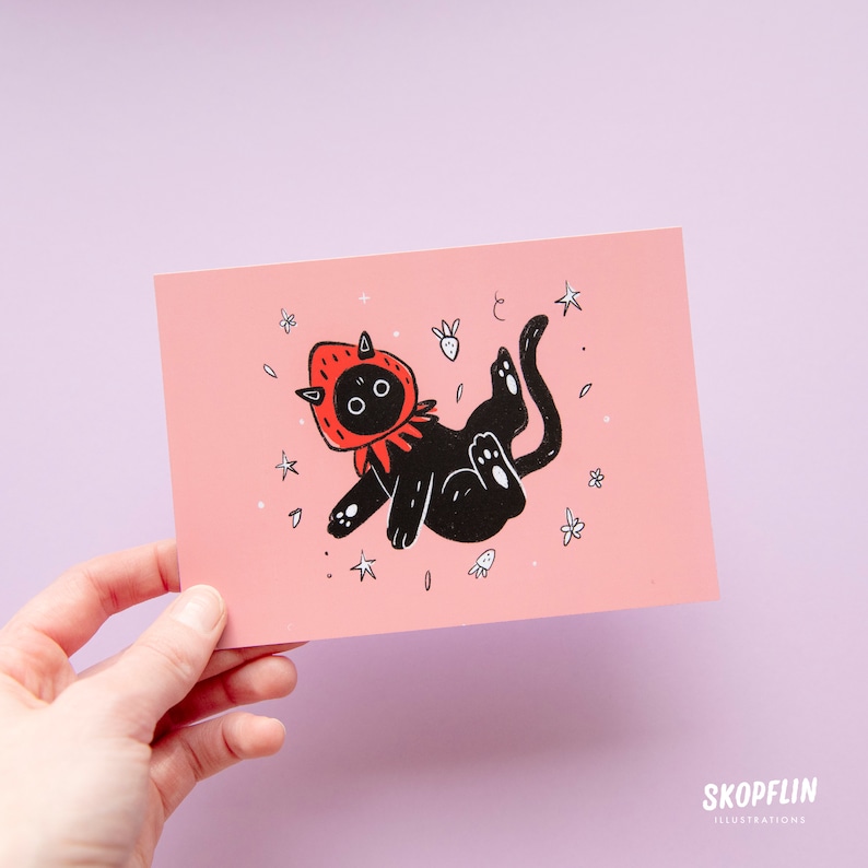 Pode incluir: Um gato preto vestindo um traje de morango vermelho flutua em um c&eacute;u rosa com estrelas e flores brancas. O gato tem olhos grandes e uma express&atilde;o fofa.