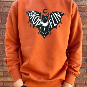 Op de afbeelding: Een oranjegekleurd sweatshirt met een zwarte vleermuis afbeelding. De vleermuis heeft een halve maan erboven en het woord "SKOPFLIN" op zijn vleugels. De vleermuis draagt een ketting met een ster hanger.