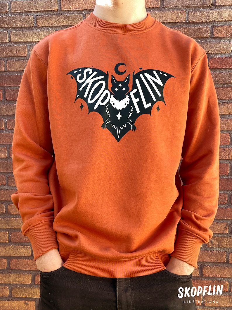 Op de afbeelding: Een oranjegekleurd sweatshirt met een zwarte vleermuis afbeelding. De vleermuis heeft een halve maan erboven en het woord "SKOPFLIN" op zijn vleugels. De vleermuis draagt een ketting met een ster hanger.