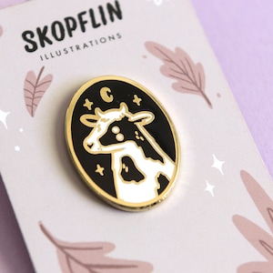 May include: A black and white enamel pin featuring a cow with a crescent moon and stars on a black background. The pin is gold-toned and has a white border. The text "C" is visible on the pin.