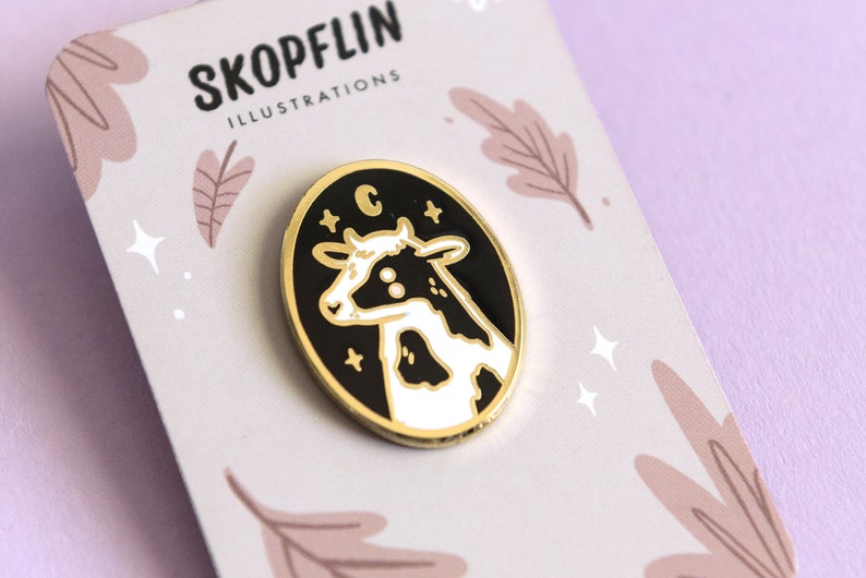 May include: A black and white enamel pin featuring a cow with a crescent moon and stars on a black background. The pin is gold-toned and has a white border. The text "C" is visible on the pin.