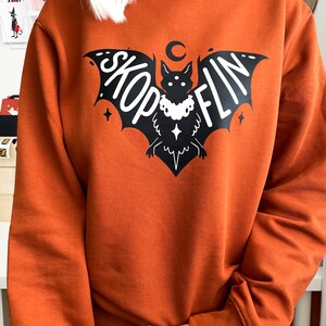 Op de afbeelding: Een oranjegekleurd sweatshirt met een zwarte vleermuis afbeelding. De vleermuis heeft een halve maan erboven en het woord "SKOPFLIN" staat op zijn vleugels.