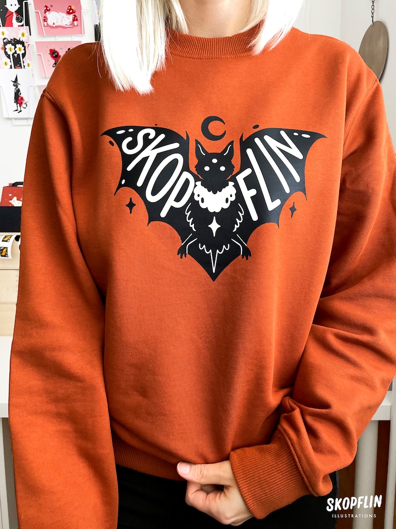 Op de afbeelding: Een oranjegekleurd sweatshirt met een zwarte vleermuis afbeelding. De vleermuis heeft een halve maan erboven en het woord "SKOPFLIN" staat op zijn vleugels.