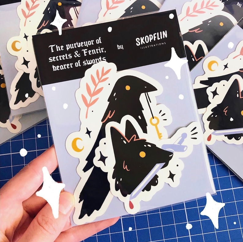 May include: A sticker featuring a black raven with a key and a wolf's head with a knife through it. The sticker has a light blue background with white stars and the text "The purveyor of secrets & Fenrir, bearer of swords" and "SKOPFLIN ILLUSTRATIONS".