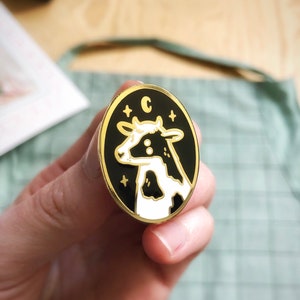 May include: A gold-colored enamel pin with a black background and a white cow with black spots. The pin has a white star on each side of the cow and the letter "C" above the cow.